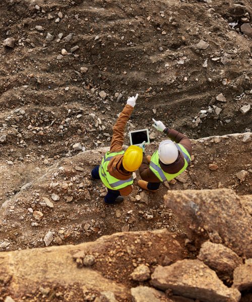 Top view shot of two industrial workers wearing reflective jackets standing on mining worksite outdoors using digital tablet, copy space