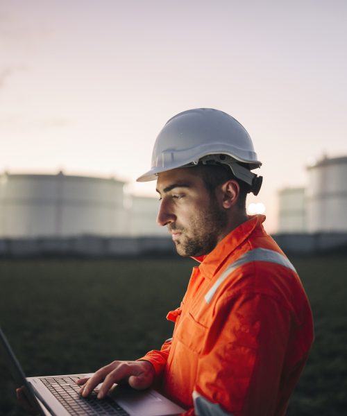 Man engineer using laptop working late night shift at petroleum oil refinery in industrial estate. Chemical engineering, fuel and power generation, petrochemical factory industry concept